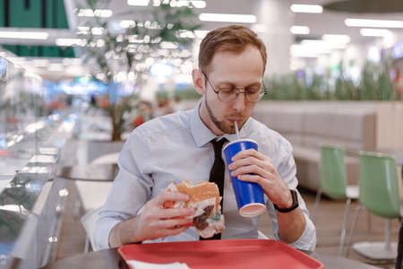 Office worker eating fast food burger and drink soda at food court in mall at lunch. Junk food at workの写真素材
