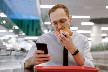 Office worker in blue shirt and black tie snacking on burger in mall, surfing the internet in a mobile while eating. Rush, social media addictionの写真素材