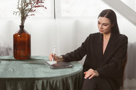 Young brunette freelancer woman designer draws sketches on a graphics tablet using a stylus. Dressed in an elegant black suit. classic interiorの写真素材