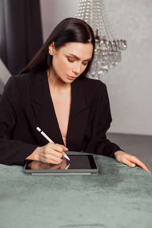 Young brunette freelancer woman designer draws sketches on a graphics tablet using a stylus. Dressed in an elegant black suit. classic interiorの写真素材