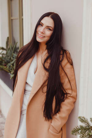 Young brunette woman in beige winter coat stands alone on the street. City street fashion, female elegant lookの写真素材