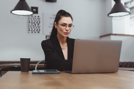 Brunette freelancer woman in black suit typing on laptop in contemporary office or cafe. chatting onlineの写真素材