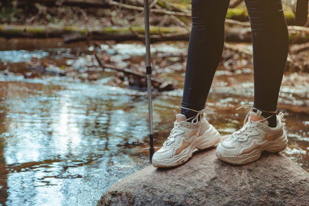 Woman Ttourist Stands in White Sneakers on a Stone in a Forest Rver. Legs and Walking stick Close upの写真素材