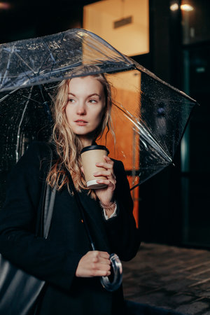 Blonde Woman in Black Coat Walking Under Transparent Umbrella and Holding Paper Cup with takeaway hot beverage in City. Cold Rainy Weather in Nightの写真素材
