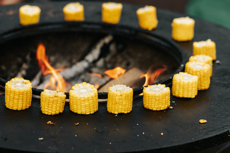 Grilled cut corn cobs on round stove with open fire outdoors, event cooking. Round steel iron fire...