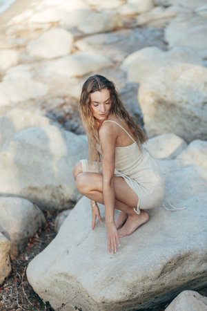 Young blonde woman in beige knitted dress sit alone on rock on beach, evening sunlight.の写真素材