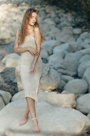Young blonde woman in beige knitted dress standing alone on rocks at beach, evening sunlightの写真素材