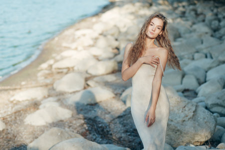 Young blonde woman in beige knitted dress standing alone on rocks at beach, evening sunlightの写真素材