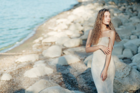 Young blonde woman in beige knitted dress standing alone on rocks at beach, evening sunlightの写真素材