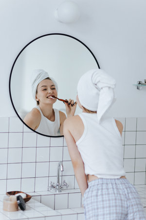 Blonde with a white towel on her head brushes her teeth in front of a mirror in a white bathroom.の写真素材