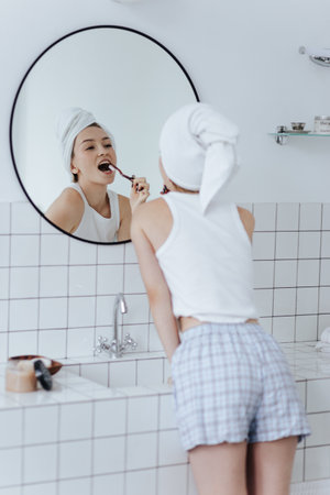 Blonde with a white towel on her head brushes her teeth in front of a mirror in a white bathroom.の写真素材