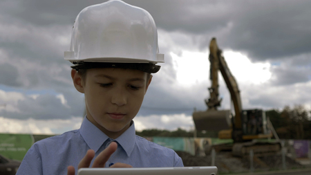 boy in a construction helmet with a tablet on the background of a combine that digs the ground, the boy is in charge of the construction projectの写真素材