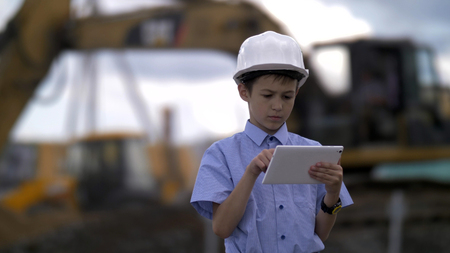 boy in a construction helmet with a tablet on the background of a combine that digs the ground, the boy is in charge of the construction projectの写真素材