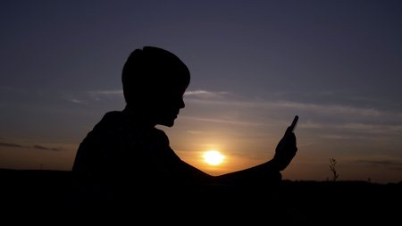 silhouette of a boy uses a tablet at sunset in the field, reads something on the tablet, outdoorsの写真素材
