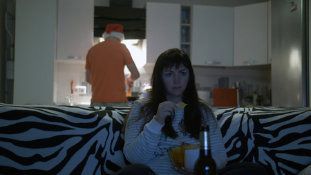 woman in front of the TV drinks beer with chips, a man prepares foodの写真素材