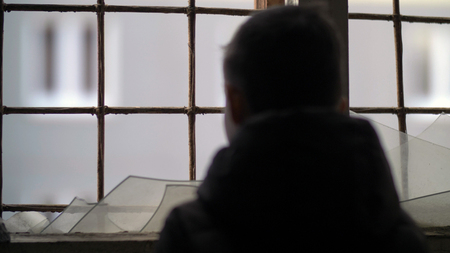 back of refugee boy stands alone near a broken window, looking out the windowの写真素材