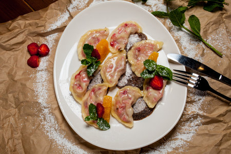 Vareniki with strawberries, mint on a large white plate, on the background of kraft paper. Next to the knife and fork - cutlery. Lenten, vegetarian menu - vareniki with cherries and strawberry sauceの写真素材