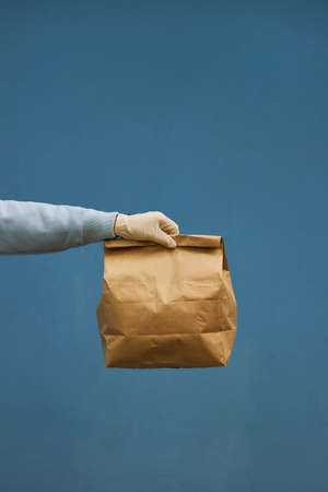 Girl courier in a medical mask and medical gloves on blue background. Delivery service in quarantine. Caucasian cute woman, delivery worker, cap and medical mask holding a grocery bag in her hands.の写真素材