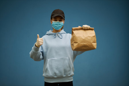 Girl courier in a medical mask and medical gloves. Delivery service in quarantine. Caucasian cute woman, delivery worker, holding a grocery bag in her hands. girl courier shows hand gesture super.の写真素材