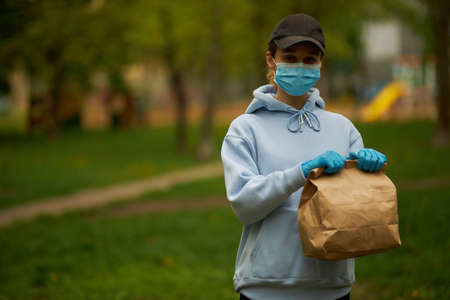 Girl courier in a medical mask and medical gloves on street. Young woman Food delivery worker. Delivery service in quarantine. Caucasian cute woman, delivery worker, holding a grocery bag in her handsの写真素材