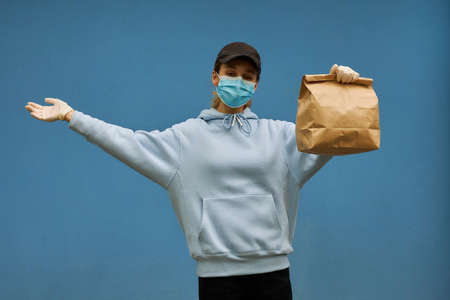 Girl courier in a medical mask and medical gloves on blue background. Delivery service in quarantine. Caucasian cute woman, delivery worker, cap and medical mask holding a grocery bag in her hands.の写真素材