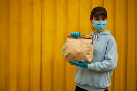 Girl courier in a medical mask and medical gloves on yellow background. Delivery service in quarantine. Caucasian cute woman, delivery worker, holding a grocery bag in her hands.の写真素材