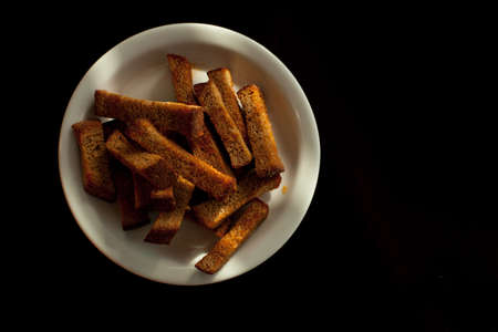 Rye croutons on a white plate isolated on a black background. Bread with croutons on a white plate. Bread gray salted croutons at white plate on a black background.の写真素材