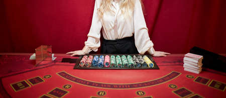 A croupier woman in a white shirt is standing at a red poker table. red Poker table in a luxury casino. Casino poker croupier womanの写真素材