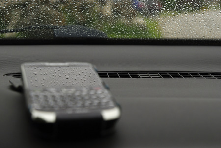 Blurred cell phone with sharp rain drops reflection in its screen. Car dashboard and windscreen with raindrops.の写真素材