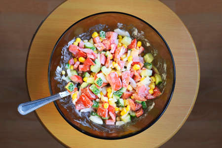 A fresh salad of chopped tomatoes, cucumbers, corn, red pepper and onions, seasoned with sour cream or mayonnaise. Vegetable glas plate on wooden background. Displaced ingredients top view.の写真素材