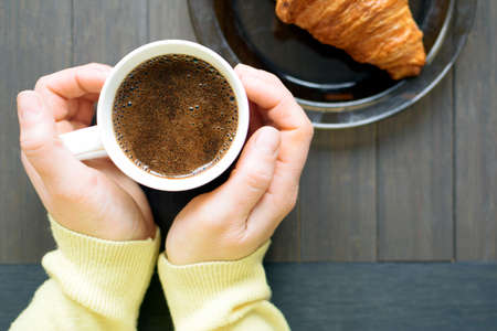 Two female hands holding cup of coffee with milk. Women's hands are warming, gently holding a cup of hot coffee, with froth. View from above on a dark wooden background.の写真素材