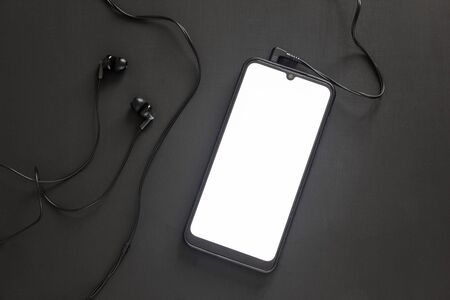 A smartphone with a white screen lies on a black background with stuck headphones.の写真素材