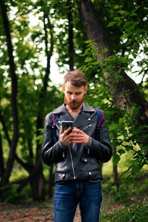 A bearded man looks thoughtfully at the phone while standing in a forest in blue waits and a black leather jacket.の写真素材