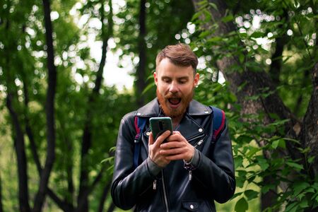 A guy with a thick beard opened his mouth wide in surprise while reading a message on a smartphone screen while standing in a dense forest.の写真素材
