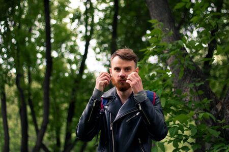Stylish guy straightens his orange mustache while standing near green trees in a black leather jacket.の写真素材