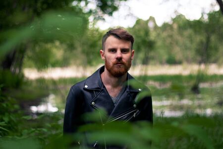 Stylish man with an orange beard and mustache stands in a green forest on a background of grass and branches.の写真素材