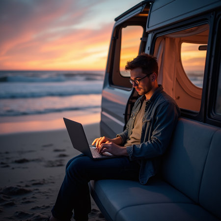 Young man with laptop sitting in camper van on beach at sunsetの素材