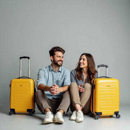 happy couple sitting on yellow travel bags and smiling at camera on gray backgroundの素材
