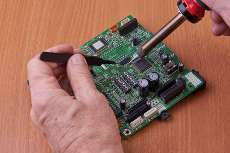 Electronic board repair component held with tweezers by an engineer over a green motherboard.