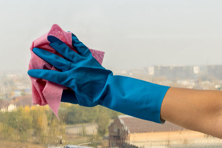 Female hand in a blue glove, cleans the window with a pink rag and detergent. Housework concept