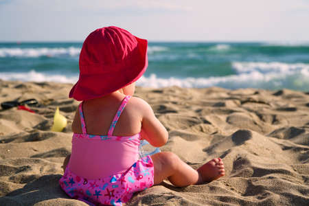 Baby in swimsuit and pink panama hat sits on beach and looks at sea on sunny dayの写真素材