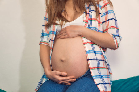A pregnant woman in jeans and a tank top with a shirt on the background of a blue sofa. Stylish...