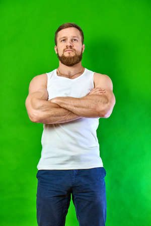 With his arms crossed over his chest, the serious man looks forward. Caucasian athlete with a beard is concentrated. Strong man on a green background in a white T-shirt and jeans.の写真素材