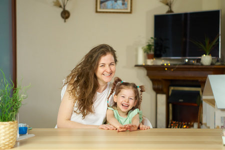 Portrait of a happy smiling family. Mom and daughter at the table look at the cameras. two sisters laugh. At home, sit in a room and laugh. Place for text. Happy childhood. woman and girl in the frameの写真素材