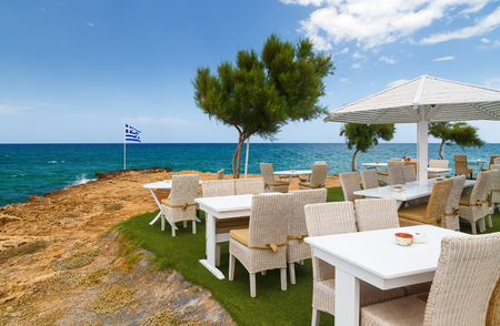 tables chairs and wooden umbrella on the rocky beach overlooking sea and Greek flag, Crete, Greeceの写真素材