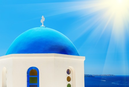 view of orthodox church blue dome and house terrace with flowers at foreground and mountains with sea at background on sunny day with sun shining in right upper corner, Oia, Santorini or Thira, Greeceの写真素材