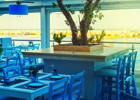 Tables and chairs at a waterfront bar with views across the harbour, Hersonissos, Crete, Greece, Europe.の写真素材