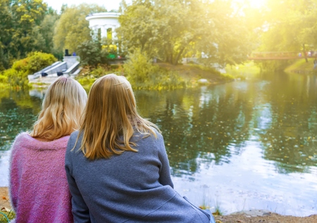 two blonde girls sit on the beach and look at the pond and the beautiful columns on otstavke, autumn city Park concept with people.の写真素材