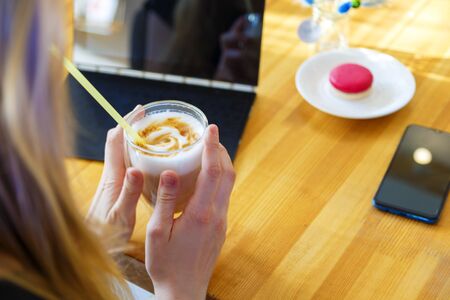 Close up female hand holding latter in glass. Back side view. Blond-haired woman freelancer or student rest after hard working day. Girl put smartphone on table, turn off laptop. Cozy cafe interiorの写真素材