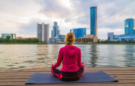 young girl meditates in a yoga pose on the shore of a city pond against the background of modern business buildings. the concept of a healthy lifestyle in the metropolisの写真素材